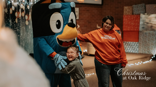 Santa Clause in a chair next to Christmas Tree with hands up waving