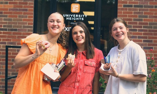 Three teen girls smile as they hold donuts up infront of church.