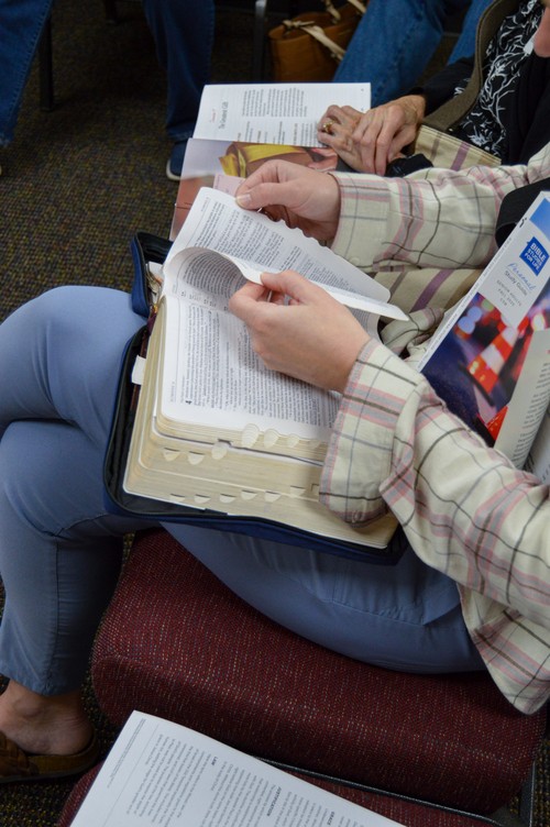 Person turning pages in an open Bible during Sunday School with study notes and booklet in their lap.