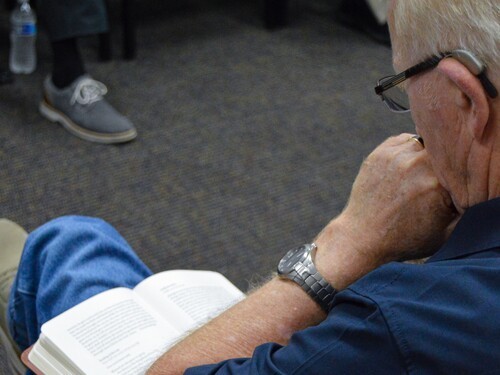 A man with a hearing aid reads his Bible in his lap with his hands clasped and propped on his chin.